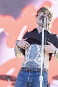 Male singer on stage, shirt lifted to reveal a tattooed chest, singing into a microphone with a bright pink backdrop behind him.