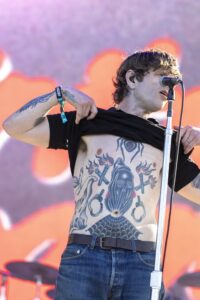 Male singer on stage shirtless with large chest tattoos, gripping a microphone stand during a live performance
