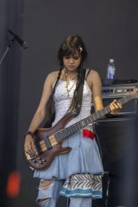 Young woman bassist performing on stage, focused on her instrument, with microphone and amp in the background.