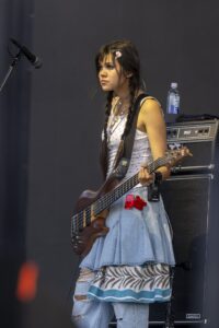 Female bassist performing on stage, playing a brown bass guitar with a strap, microphone to her left, and an amplifier in the background.