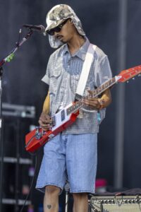 Male musician wearing a camouflage hat and sunglasses, playing a red rectangular guitar on stage with a microphone nearby.
