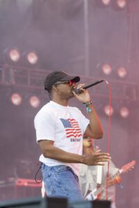 Male singer on a smoky stage wearing a white T-shirt with an American flag graphic, blue jeans, a black cap, and red sunglasses, singing into a handheld microphone with a red cable.