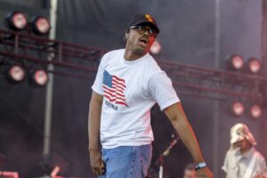 Male performer on stage wearing a white T-shirt with an American flag graphic, jeans, glasses, and a cap, mid-song during a live show.