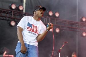 Male singer on stage wearing a white T-shirt with an American flag graphic, jeans, cap and sunglasses, holding a red microphone cord and singing.