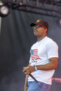 Male performer on stage wearing a black cap, sunglasses, white USA flag T-shirt, and jeans, holding a microphone.