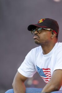 Man wearing a black cap with a Porsche logo, mirrored sunglasses, and a white T‑shirt with an American flag, looking to the left outdoors.