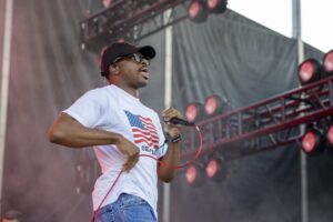 Male vocalist on stage wearing a white T-shirt with an American flag, cap, and sunglasses while holding a microphone with a red cord.