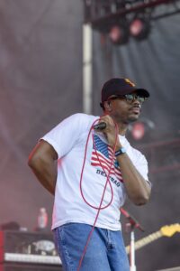 Male singer on stage wearing a white American flag T-shirt, cap, and sunglasses, holding a red microphone cord to his mouth.