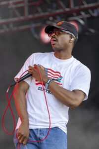 Male singer on stage, wearing a black cap and red sunglasses, holding a microphone with red cables looping around him.