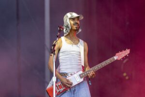 Male guitarist performing on stage in a white tank top and camouflage hat, playing a red and white electric guitar.