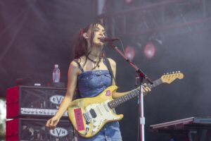 Female guitarist singing into a microphone on stage, playing a yellow Fender-style electric guitar.