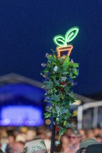 Neon green leaf-shaped sign atop a plant-covered pole at night, with a blurred crowd in the background.