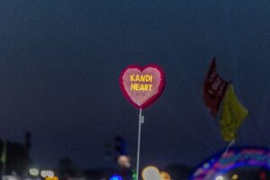 Pink heart-shaped balloon on a string with 'KANDI HEART' in yellow letters against a dark night sky, festival lights in the background.