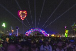 Nighttime festival scene with a large colorful Bonnaroo arch, laser beams shooting into the sky, and a dense crowd in silhouettes at the foreground.
