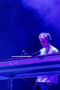 DJ wearing headphones stands at a mixing console under blue stage lights, focused on equipment.