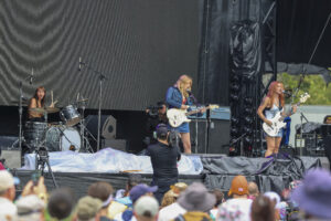 Live outdoor concert band performing on stage: drummer left, guitarist center, bassist right, with a crowd in the foreground.