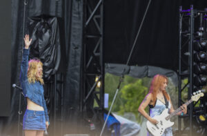 Young female singer in a denim outfit raises her hand on stage during a concert, with a guitarist playing a white bass nearby.