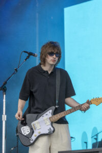 Male guitarist wearing sunglasses and a black polo plays a black electric guitar on a blue stage backdrop.