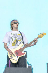 Male guitarist wearing sunglasses and a graphic white T-shirt, playing a cream-colored bass guitar on a blue stage backdrop.