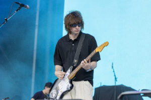 Male guitarist wearing sunglasses and a black polo plays a white electric guitar on a blue stage backdrop with a microphone nearby