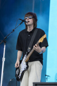 Male guitarist singing into a microphone on stage, wearing sunglasses and a black polo, playing a white Stratocaster against a blue backdrop.