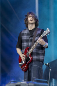 Male guitarist with long hair and beard, wearing a plaid vest, playing a red Gibson SG on stage.