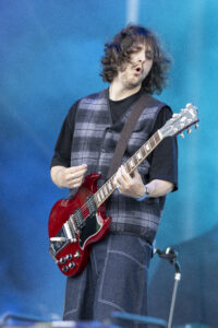 Guitarist performing on stage, playing a red electric guitar against a blue background.