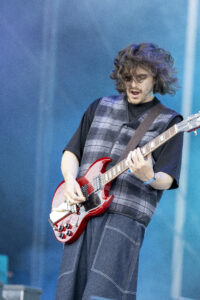 Guitarist with long hair playing a red electric guitar on a blue stage backdrop, wearing a plaid vest and black shirt.