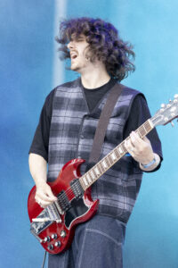 Guitarist with long curly hair playing a red electric guitar on a blue stage backdrop, wearing a plaid vest.