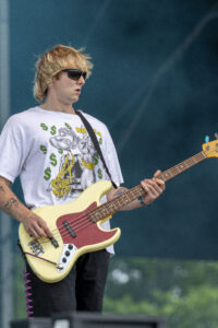 Male bass guitarist wearing sunglasses and a graphic white T-shirt, playing a cream-colored bass on stage.