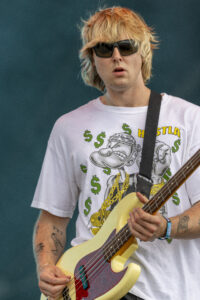 Musician with blond hair and sunglasses playing a cream-colored bass guitar on stage, wearing a white graphic tee and strap across chest.