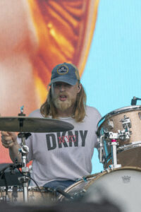 Drummer with long hair and a blue Corona cap playing a drum kit on a colorful stage backdrop.