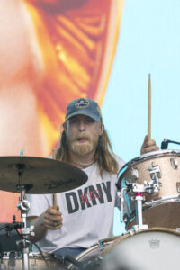 Drummer with long hair and beard wearing a blue Corona cap playing a drum kit on a stage, holding drumsticks against a bright orange-blue backdrop.