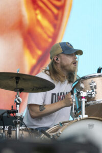 Drummer with long blond hair and a blue-tan cap playing a drum kit on stage, colorful background behind him.