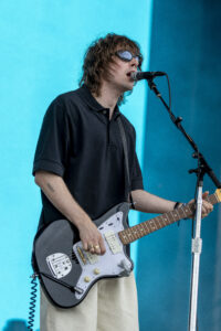 Musician singing into a microphone while playing a black electric guitar on a blue stage backdrop.