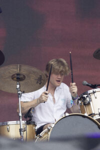 Drummer in a white shirt playing a drum kit on a stage with cymbals and a microphone nearby.