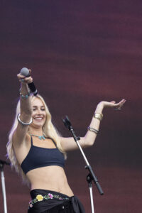 Female singer on stage raises a microphone, wearing a navy crop top and a floral belt with turquoise jewelry, against a purple sunset backdrop.