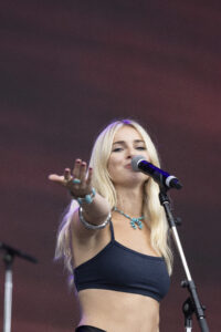 Female singer with long blonde hair reaches toward the audience while singing into a microphone on stage menagerie of purple lights behind her are visible in the background.
