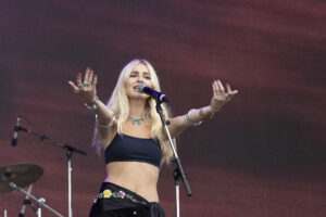 Female singer on stage with microphone, arms outstretched in performance, wearing a black crop top and turquoise jewelry.