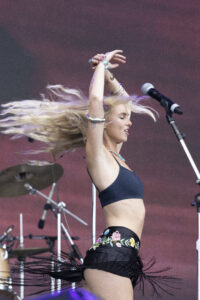 Female performer on stage with arms raised above head, hair flying, wearing a navy crop top and floral shorts, microphone nearby and drum kit in background.