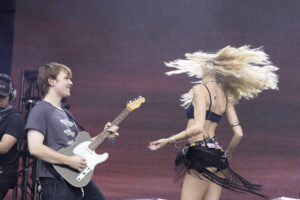 Male guitarist playing a white Fender-style electric guitar on stage, smiling as a female dancer with long blonde hair moves energetically nearby on a darkened backdrop in a live performance.