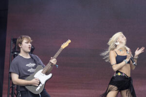 Two musicians performing on stage: a male guitarist on the left and a female singer with a microphone on the right against a warm gradient backdrop.