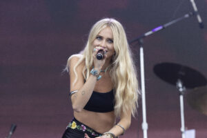 Blonde female singer on stage holding a microphone, wearing a black crop top and turquoise jewelry, performing with drums in the background