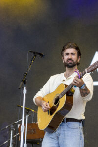 Male singer-guitarist performing on stage with an acoustic guitar and microphone in front of him, wearing a light shirt and jeans