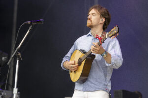 Male musician performing live, playing an acoustic guitar on stage with a microphone stand nearby, eyes closed and wearing a light striped shirt.