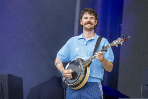 Smiling man with a mustache playing a banjo on stage, wearing a light blue polo shirt against a blue backdrop.