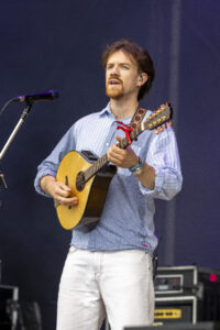 Male guitarist performing on stage, singing into a microphone while playing an acoustic guitar with a red capo on the neck.