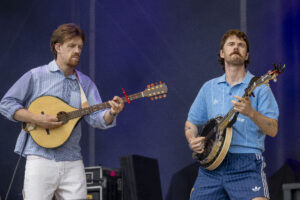 Two male musicians performing on stage with guitars; left guitarist in a striped shirt, right guitarist in a blue polo with a mustache.