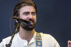 Male singer with a beard performing on stage into a microphone, wearing a light shirt and a colorful patterned guitar strap.