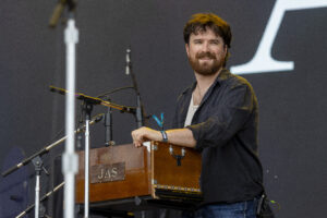 Musician with a beard and dark shirt playing a wooden resonator guitar on stage, with microphones nearby.